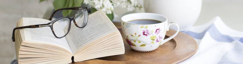 Bild mit einem Buch einer Brille darauf und einer Teetasse nebenbei auf einem Holztablett